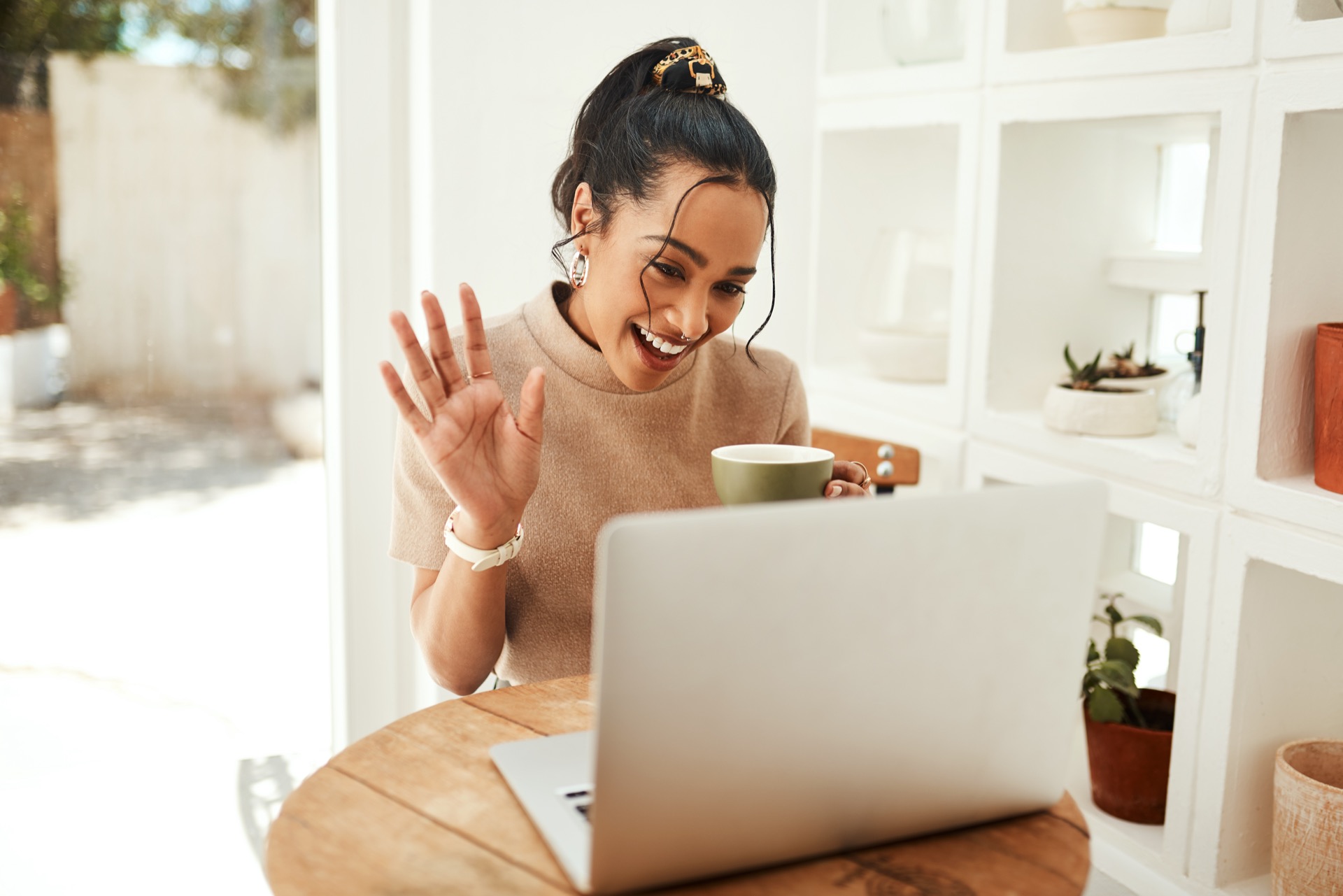 Young woman sitting and waving to people in a video call on her laptop while enjoying a cup of coffee
